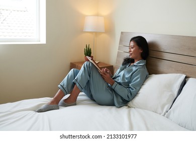 Smiling young woman in silk pajamas sitting on bed and reading ebookk on tablet computer