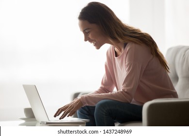 Smiling young woman sit on couch using laptop look at screen feel excited reading good news online, overjoyed girl relax on sofa laugh typing messaging on computer, or shopping on internet