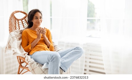 Smiling young woman sits in a wicker armchair indoors, sipping coffee, looking aside at empty space for text. Arabian lady enjoys weekend morning with hot drink at comfort of home. Panorama - Powered by Shutterstock