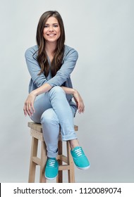 Smiling young woman sitting on stool with crossed legs. Isolated studio portrait on gray back.