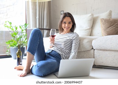 Smiling young woman sitting on floor with laptop computer and chating with friends, drinking wine