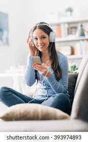 Smiling young woman sitting on the sofa at home and listening to music, she is wearing white headphones