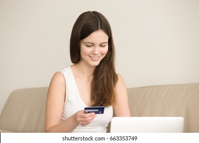 Smiling Young Woman Sitting On Sofa With Laptop And Looking On Credit Card In Her Hand. Happy Lady Making Purchases In Internet Shop, Teen Girl Using Online Banking Service, Checking Account Balance