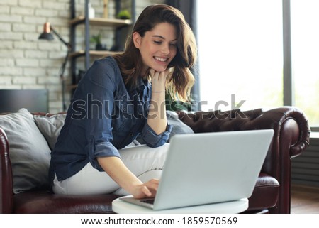Smiling young woman sitting on sofa with laptop computer and chating with friends.