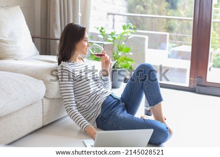 Smiling young woman sitting on floor with laptop computer and chating with friends, drinking wine