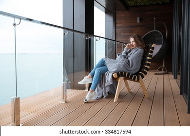 Smiling young woman sitting wrapped in knitted coverlet on the glass balcony