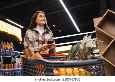 Smiling Young Woman With Smartphone In Supermarket. Pretty Girl With Shopping Cart In Supermarket Using Mobile Phone
