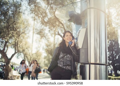 Smiling Young Woman Speaking On A Public Payphone Outside.Happy Expression.Dialing Local Number In Foreign Country,low Cost Calls For International Dials.Coin Or Card Public Phone Used By Tourist