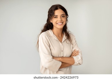 Smiling young woman standing confidently with arms crossed over light background, happy beautiful female manager wearing blouse exuding professionalism, looking at camera, copy space - Powered by Shutterstock