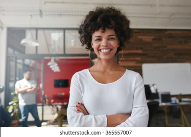 Smiling young woman standing with her arms crossed and looking at camera. She is standing in a modern office with her colleagues in the background. - Shutterstock ID 365557007