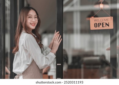 A smiling young woman stands by a glass door, welcoming customers with an "OPEN" sign in a cozy café setting. - Powered by Shutterstock