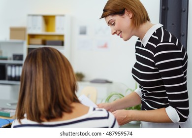 Smiling Young Woman In Striped Shirt Standing Near Seated Female Coworker In Office Setting