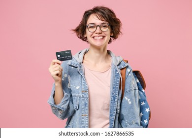 Smiling young woman student in denim clothes glasses backpack posing isolated on pastel pink background. Education in high school university college concept. Mock up copy space. Hold credit bank card