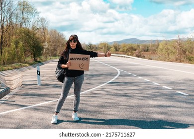 Smiling young woman in sunglasses and cap holding a cardboard sign with text anywhere. Copy space. The concept of local traveling and hitchhiking.