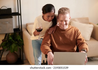 Smiling Young Woman Supporting Her Boyfriend In Wheelchair Working On Laptop, Stidying Or Searching For New Job