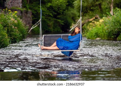 A Smiling Young Woman Swings On A Rope Swing Across A Fast-flowing River