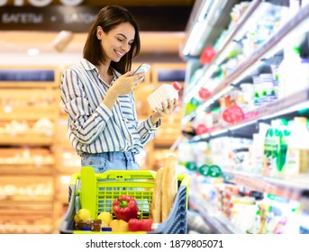 Smiling Young Woman Taking Dairy Products From Shelf In The Supermarket, Holding Bottle And Smartphone, Scanning Bar Code On Product Through Mobile Phone, Walking With Trolley Cart