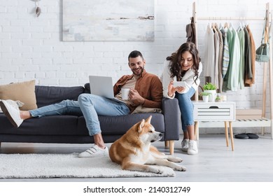 smiling young woman taking photo of akita inu dog near man using laptop in living room