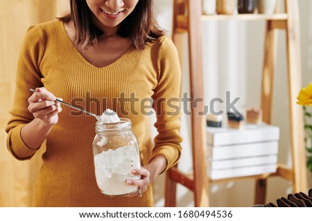 Smiling young woman taking spoon of soda lye from glass jar when making fragrant soap at home