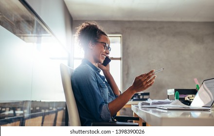 Smiling young woman talking on phone with a laptop in front. Businesswoman in conversation over phone at office. - Powered by Shutterstock