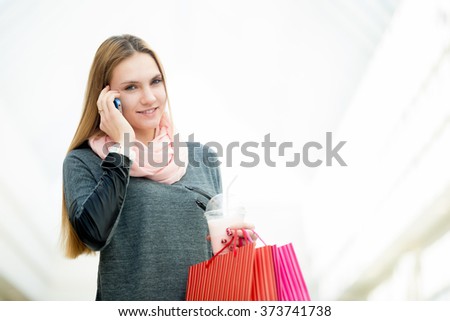 Smiling young woman talking on cellphone while holding paper bags and strawberry cocktail in shopping center, copyspace