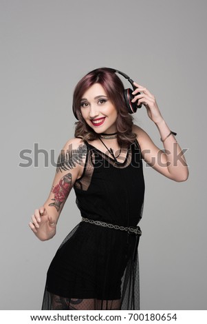 Smiling young woman with tattoos dancing while listening to music in big headphones and looking at camera