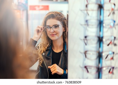 Smiling Young Woman Trying On Glasses On Mirror In Optician.