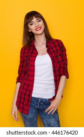 Smiling Young Woman In Unbuttoned Red Lumberjack Shirt And Jeans. Three Quarter Length Studio Shot On Yellow Background.