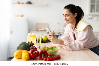 Smiling young woman using cellphone while cooking dinner in kitchen at home, cheerful casual female leaning on table, reading text messages on her cell phone and smiling, free copy space - Powered by Shutterstock