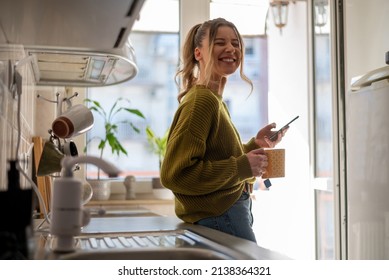 Smiling young woman using her smartphone while drinking a cup of coffee in the kitchen at home. In the morning concept. Smiling woman reading phone message. - Powered by Shutterstock