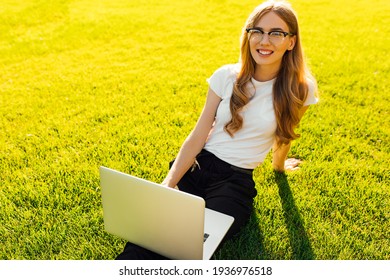 Smiling young woman using laptop sitting on green grass in park outdoors, beautiful girl shopping or chatting online on social network, freelancer working on computer project - Powered by Shutterstock