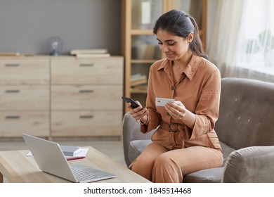 Smiling Young Woman Using Mobile Phone And Credit Card For Online Payment While Sitting On Sofa In Front Of The Laptop