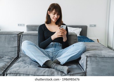 Smiling young woman using mobile phone while sitting on a couch at home - Shutterstock ID 1986985676