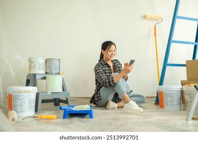 Smiling young woman using mobile phone, taking a break from painting during a home renovation project. - Powered by Shutterstock