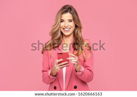 Smiling Young Woman Using Mobile Phone, looking at camera, wearing fashionable jacket. Pink studio background. Smart businesswoman. Hapy student.