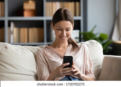 Smiling young woman using phone, sitting on couch at home, looking at smartphone screen, beautiful female chatting in social network or shopping online, spending leisure time with mobile device