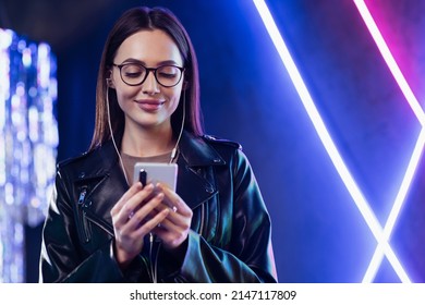 Smiling young woman using smartphone in a night club. Trendy girl wearing glasses looking at mobile phone. People, mobile apps, modern lifestyle, technology concept