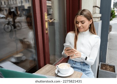 smiling young woman using smartphone near cup of cappuccino while sitting in cafe in Vienna - Powered by Shutterstock