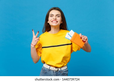Smiling young woman in vivid casual clothes looking camera, showing victory sign, holding passport, boarding pass ticket isolated on blue wall background. People lifestyle concept. Mock up copy space