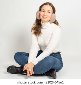 Smiling Young Woman In Warm White Jumper With Fur Earmuffs Isolated On White Sitting On The Floor With Crossed Legs In Jeans