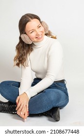 Smiling Young Woman In Warm White Jumper With Fur Earmuffs Isolated On White Sitting On The Floor With Crossed Legs In Jeans