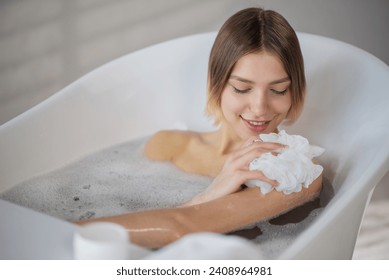 Smiling young woman washing with soapy sponge in bath in morning. View from above of happy female scrubbing body skin with washcloth, while soaking in bathtub. Concept of bathing process. - Powered by Shutterstock