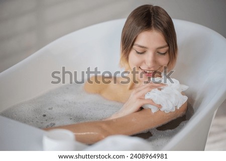 Smiling young woman washing with soapy sponge in bath in morning. View from above of happy female scrubbing body skin with washcloth, while soaking in bathtub. Concept of bathing process.