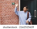 Smiling young woman waving with a friendly cheerful smile to her new neighbours. Girl leaves the house closing the door and waving her hand. Girl wear blue sweater and brown bag, meeting friends.