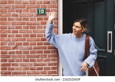 Smiling young woman waving with a friendly cheerful smile to her new neighbours. Girl leaves the house closing the door and waving her hand. Girl wear blue sweater and brown bag, meeting friends.