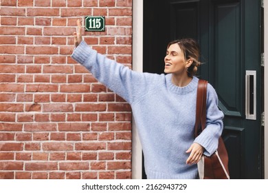 Smiling Young Woman Waving With A Friendly Cheerful Smile To Her New Neighbours. Girl Leaves The House Closing The Door And Waving Her Hand. Girl Wear Blue Sweater And Brown Bag, Meeting Friends.