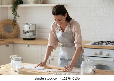 Smiling young woman wearing apron rolling out dough with rolling pin in hands, happy female preparing breakfast, baker cooking cake or cookies in kitchen at home, standing at table - Powered by Shutterstock