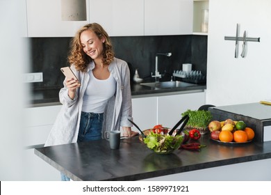 Smiling young woman wearing casual clothes using mobile phone while leaning on a kitchen table - Powered by Shutterstock