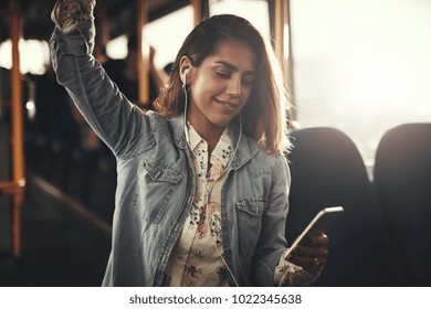 Smiling young woman wearing earphones standing by herself on a bus listening to music on a smartphone 