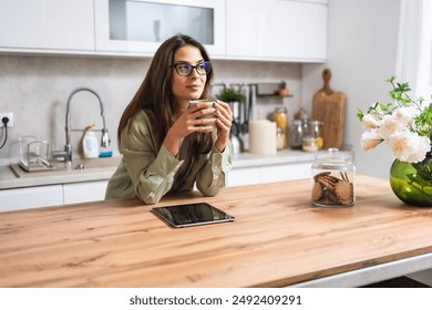 Smiling young woman wearing glasses drinking coffee and using tablet computer while sitting in kitchen. Businesswoman reading emails at morning in her home. Remote work business female - Powered by Shutterstock